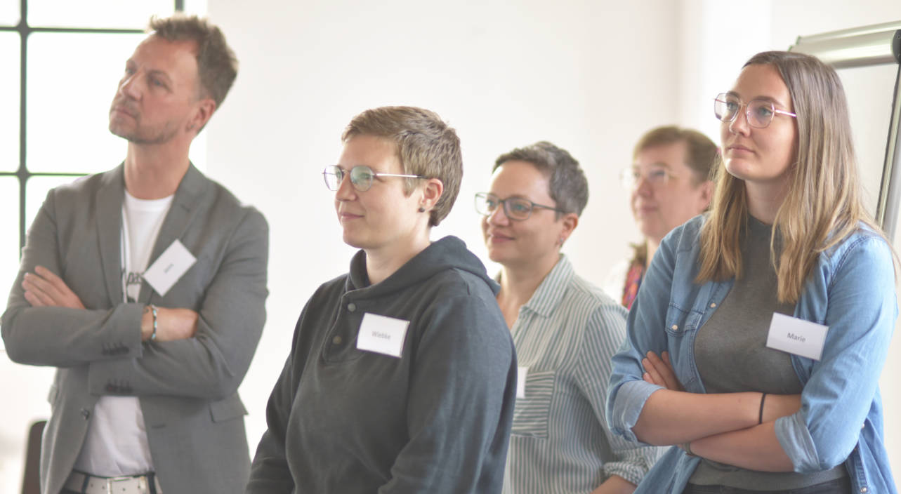 Auf dem Bild sind drei Personen zu sehen, die gemeinsam an einer Aufgabe arbeiten. Im Mittelpunkt steht eine Frau mit Brille und braunen Haaren. Sie schaut konzentriert auf die Hände der anderen Personen. Alle halten gemeinsam ein dünnes, gelbes Stäbchen und bauen offenbar etwas Kleines. Die Stimmung wirkt ruhig und fokussiert. Diese Szene zeigt eine Übung, wie sie in einer Ausbildung als Trainer*in vorkommt.